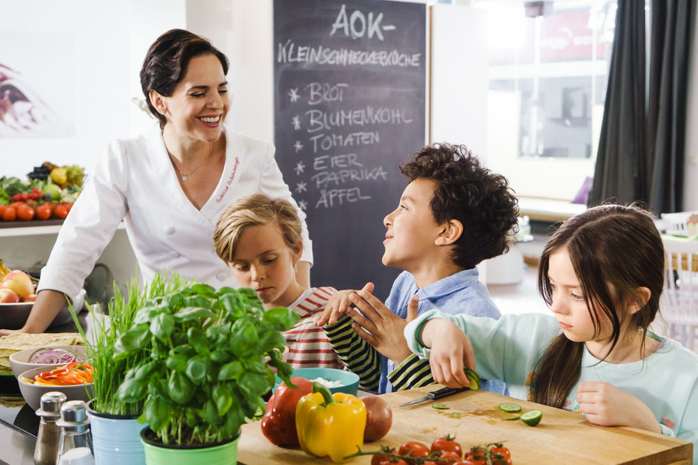 Köchin und Kinder schneiden Gemüse in einer Küche mit Tafel im Hintergrund und frischem Basilikum.